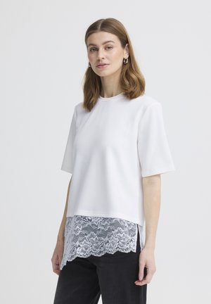 Femme aux cheveux bruns mi-longs portant un haut blanc à manches courtes avec un ourlet en dentelle superposé et un pantalon noir, debout devant un fond clair uni.