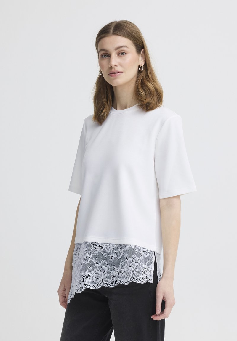 Femme aux cheveux bruns mi-longs portant un haut blanc à manches courtes avec un ourlet en dentelle superposé et un pantalon noir, debout devant un fond clair uni.