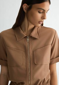 Jumpsuit corto marrone a maniche corte con zip frontale, due tasche sul petto e vita arricciata. Realizzato in tessuto leggero e morbido.