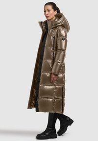 Long, shiny puffer coat in metallic bronze with a hood, black accents, zippered pockets, and a quilted pattern. Paired with black ankle boots.