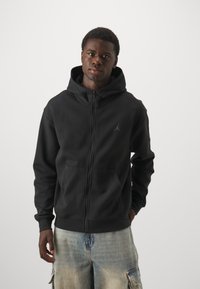 Sort zip-up hættetrøje med lomme foran og snøre. Har et diskret sort logo på venstre bryst. Bæres med en lys denimshorts.