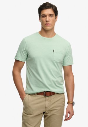 Hombre con camiseta de manga corta color verde claro con bolsillo, pantalones beige, cinturón de cuero marrón y reloj de pulsera, de pie con una mano en el bolsillo.