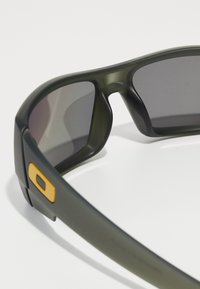 Ochelari de soare din plastic verde cu lentile închise, având un cadru gros și un accent galben pe lateral. Designul subliniază o estetică sportivă.