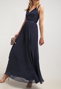 Žena v bezramínkových námořnických modrých šatech až na zem s plisovaným živůtkem, držící béžovou kabelku a obutá v béžových sandálech na vysokém podpatku.