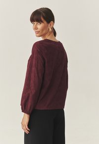 Burgundy pitkähihaine paita, jossa on teksturoitu kangas, pyöreä pääntie ja frillatut hihat. Kulta-väriset kolmion muotoiset korvakorut täydentävät ilmettä.