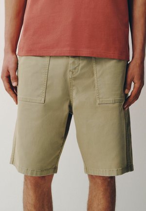 Mann trägt beige knielange Shorts mit großen aufgesetzten Taschen vorne und ein rostfarbenes T-Shirt, steht mit entspannten Armen.