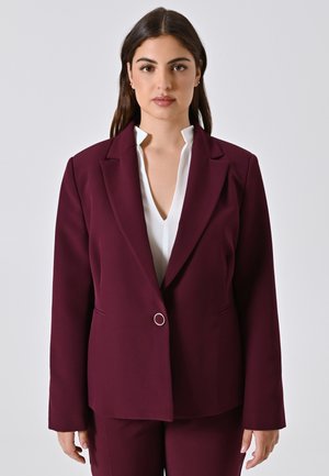 Femme aux longs cheveux noirs portant un costume bordeaux avec une veste à un bouton et un chemisier blanc à col en V, se tenant devant un fond uni.
