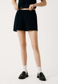 Zwarte shorts met een losse pasvorm, elastische tailleband, zijzakken en een zachte textuur, gecombineerd met zwarte loafers en witte sokken.