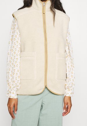 Veste sans manches en polaire beige avec fermeture éclair et deux poches avant portée sur un chemisier blanc à manches longues avec un petit imprimé floral jaune.