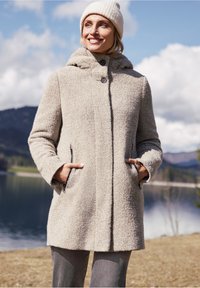 Lichtbeige wollen jas met een capuchon, voorzien van een textuur, twee zijzakken en een knoopsluiting. Gecombineerd met een crèmekleurige gebreide muts.