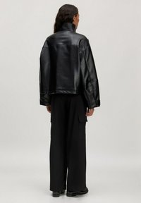 Schwarze Kunstlederjacke mit hohem Kragen, übergroßem Design und fallenden Schultern, kombiniert mit weiten schwarzen Hosen und flachen Schuhen.