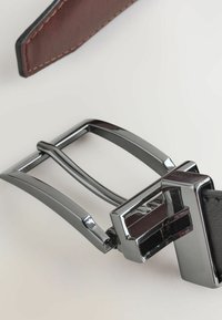 Gros plan d'une ceinture en cuir noir avec une boucle rectangulaire en argent brillant et une pointe en cuir marron sur une surface claire.