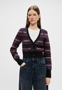 Crni i ljubičasti kardigan s uzorkom i gumbima, s natpisom "TOMMY JEANS". Materijal je meka pletenina; skraćeni dizajn s dugim rukavima.