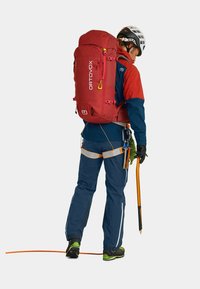 Red Ortovox backpack with adjustable straps, attached carabiner, and a solid design, worn by a person in blue and orange outdoor gear.