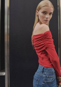 Crvena off-shoulder majica s detaljima nabrane tkanine u kombinaciji s plavim trapericama visokog struka. Glatka tekstura, prilagođen dizajn i svakodnevni stil.