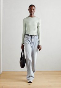 Lys grøn langermet topp, lys blå høydejusterbar jeans, svart håndpose, sølv sportsko, glatt tekstur, står på tregulv.