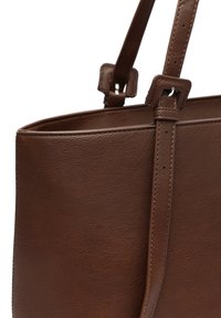Brązowa skórzana torba typu tote o gładkiej teksturze, z regulowanymi paskami i minimalnym detalem w postaci szycia. Ma prostokątny kształt i otwartą górę.