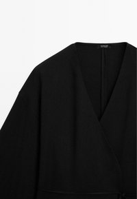 Indumento nero a forma di pacchetto con scollo a V, texture morbida e maniche ampie. Presenta una cucitura discreta lungo i bordi e l'apertura sul retro.