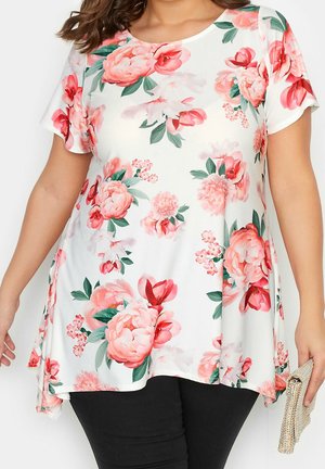 Biała tunika z krótkim rękawem z dużym różowym nadrukiem kwiatowym, noszona z czarnymi spodniami, trzymająca w prawej ręce beżową, fakturowaną kopertówkę.