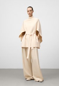 Beige oversized top met hoge kraag en zijsplitten, met een ceintuur op de taille. Gecombineerd met lichtbeige wijde pantalon en zachte, ronde schoenen.