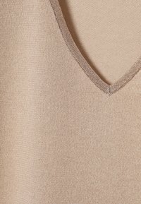 Beige neulottu yläosa V-pääntiellä, jossa on sileä rakenne ja hienovarainen kiilto. Reunoissa on hieno metallityylinen koriste.