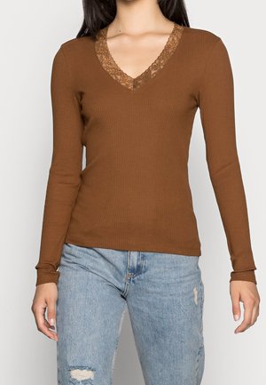 Kvinde iført en brun ribstrikket bluse med lange ærmer og blonder ved V-udskæringen, kombineret med lyseblå distressed jeans.