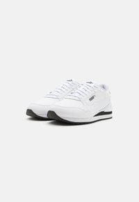 Des chaussures de sport blanches avec une tige synthétique lisse, un design à lacets, une semelle en caoutchouc foncé et un branding discret sur le côté et la languette.