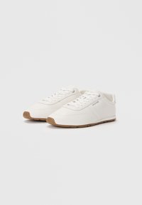 Adidași albi cu un upper din textile netede, vârf rotund și o talpă din cauciuc bej. Au șireturi și un branding subtil pe lateral.