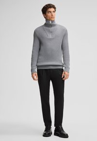 Maglione grigio a maglia con colletto alto e mezza zip, abbinato a pantaloni neri e scarpe nere. Il maglione presenta una texture a coste.