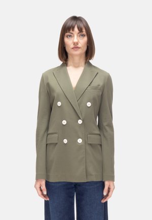 Donna che indossa un blazer doppiopetto verde oliva con bottoni bianchi e jeans blu, in piedi contro uno sfondo chiaro e uniforme.