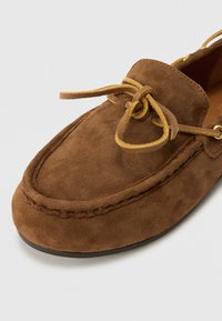 Scarpe mocassino in suede marrone con punta arrotondata, lacci decorativi e suola cucita. La superficie presenta una finitura morbida e testurizzata.