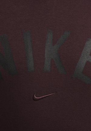 Maroon athletic shirt with large black "NIKE" lettering. Features a small embroidered black Nike swoosh. Made from smooth, stretchy fabric.