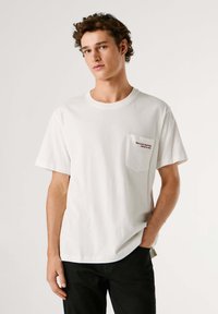 Camiseta blanca de algodón con un bolsillo delantero y texto en rojo. Corte clásico con mangas cortas y cuello redondo, combinada con pantalones oscuros.