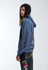 Navy hoodie featuring graphic prints of an octopus and text on sleeves, made of soft fabric with ribbed cuffs and hem. Paired with black jeans.