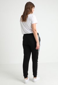 Melnas bikses ar baltām sānu svītrām, elastīgu jostasvietu un sašaurinātiem galiem, apvienotas ar pieguļošu baltu T-kreklu, redzamas no aizmugures.