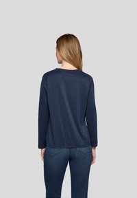 Frau mit glatten, hellbraunen Haaren, die ein marineblaues Langarmoberteil und dunkelblaue Jeans trägt, mit dem Rücken zu einem schlichten Hintergrund gerichtet.