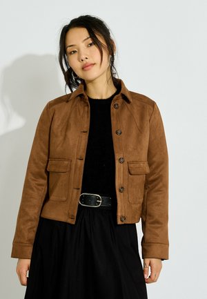 Mujer con cabello oscuro que lleva una chaqueta marrón abotonada sobre una blusa negra y falda con cinturón negro, de pie frente a un fondo liso y claro.