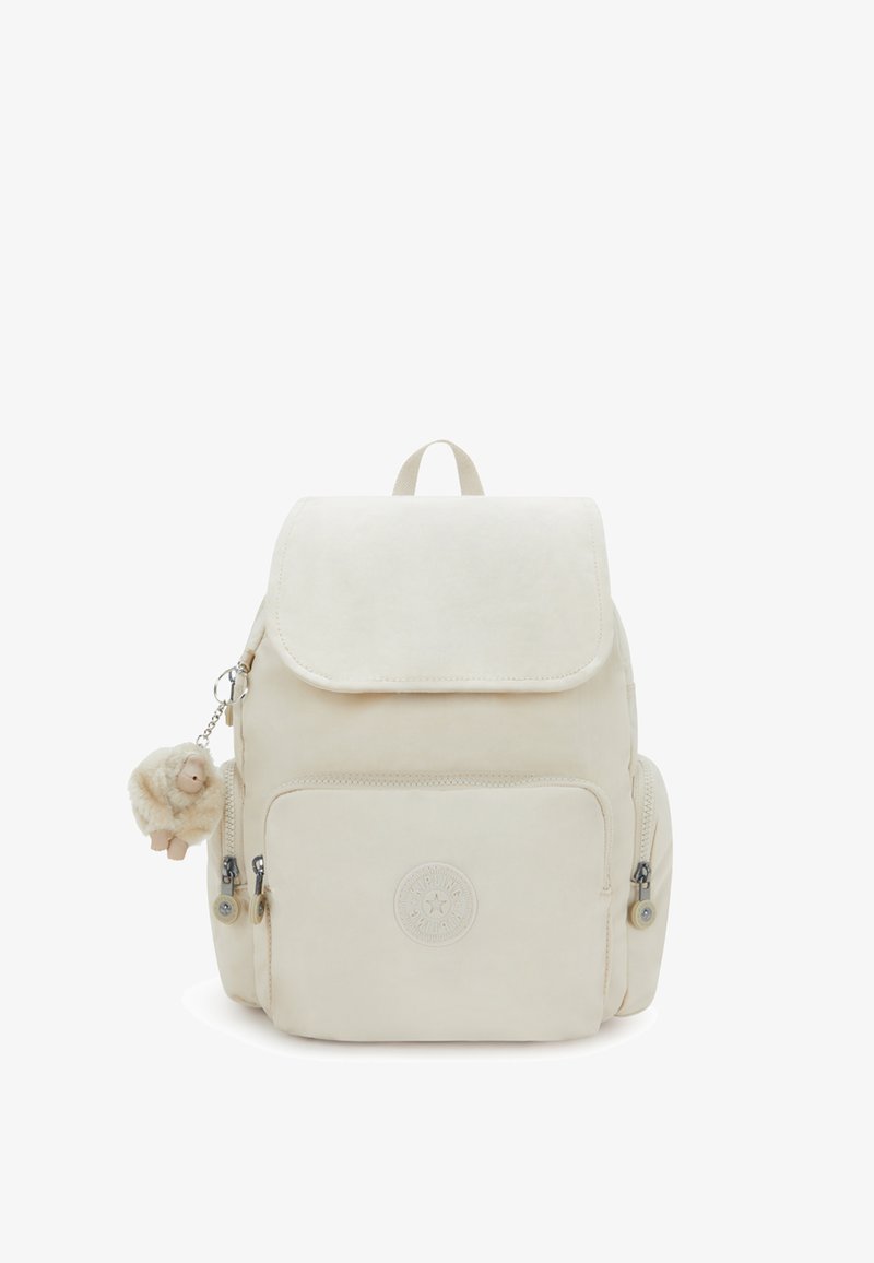 Mochila beige con cierre de solapa, que presenta un logo redondo, un bolsillo con cremallera en la parte frontal, bolsillos laterales y un llavero de adorno esponjoso. Textura suave.