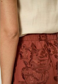 Gonna marrone ricamata con motivi floreali, vita arricciata e un top beige chiaro. Tessuto strutturato visibile, che mostra dettagli intricati.