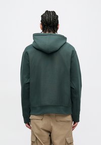 Sudadera verde con acabado texturizado, que presenta una capucha espaciosa y un dobladillo acanalado. Combinada con pantalones cargo caqui, mostrando detalles de los bolsillos.