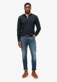 Suéter de marina con cremallera de un cuarto, cuello gris, jeans azules y botas de cuero marrón. Tejido suave con un ajuste casual y detalles de diseño mínimos.
