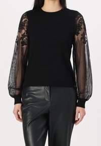 Pullover nero con corpo slim e maniche in pizzo nero trasparente plissettate con ricami floreali. Abbinato a pantaloni in pelle nera lucida.