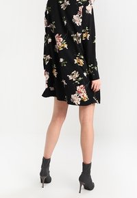Schwarz-blumiges Kleid mit langen Ärmeln, das mit rosa und gelben Blumen verziert ist. Glatte Stoffqualität mit fließender Silhouette, kombiniert mit Stiefeln bis Knöcheln.