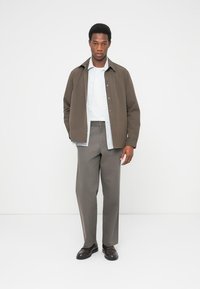 Καφέ σακάκι με κουμπιά πάνω από ένα ανοιχτό μπλε πουκάμισο με ρίγες, γκρι παντελόνια και μαύρα παπούτσια· λείο ύφασμα και εφαρμοστή γραμμή.