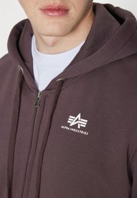 Sudadera con cremallera marrón hecha de algodón, con un logo blanco en el lado izquierdo del pecho, capucha con cordón y tela texturizada.