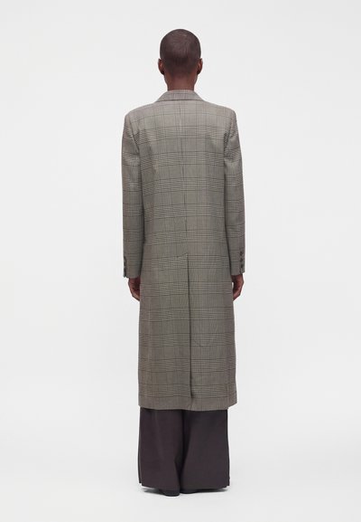 Personne aux cheveux courts portant un long manteau à carreaux et un large pantalon sombre, de dos, sur un fond clair uni.