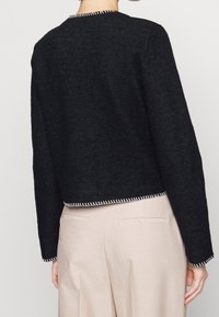 Czarny krótki sweter z teksturowanej tkaniny, z białymi kontrastującymi szwami wzdłuż dekoltu i dołu. Zestawiony z jasnobeżowymi spodniami.