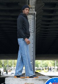 Svart hettegenser, lyseblå vide jeans og svarte sko med oransje såler. Står på en betongoverflate nær en metallstruktur.