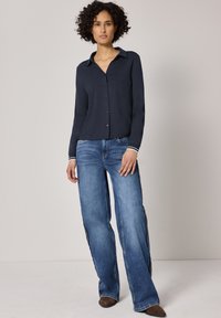 Femme aux cheveux bouclés portant une chemise bleu marine boutonnée, un jean large bleu avec des bandes latérales, et des chaussures marron pointues, debout sur un fond uni.