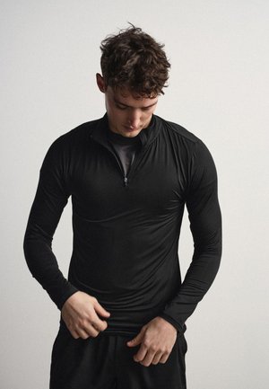 Joven ajustándose la camiseta deportiva negra de manga larga con cremallera frontal, de pie frente a un fondo blanco liso.